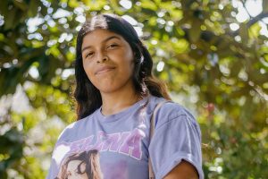 A student in a lavender Selena t-shirt posts outside in front of greenery