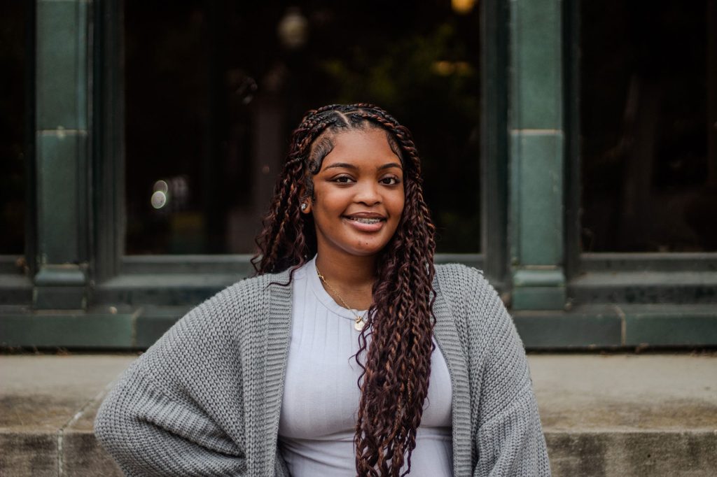 A girl with braids wearing a grey sweater and a white t shirt stands in front of a building with green framing on the outside.