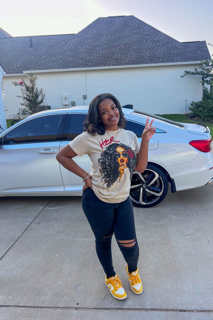 A young lady smiles for a photo on first day of school standing in front of a white car