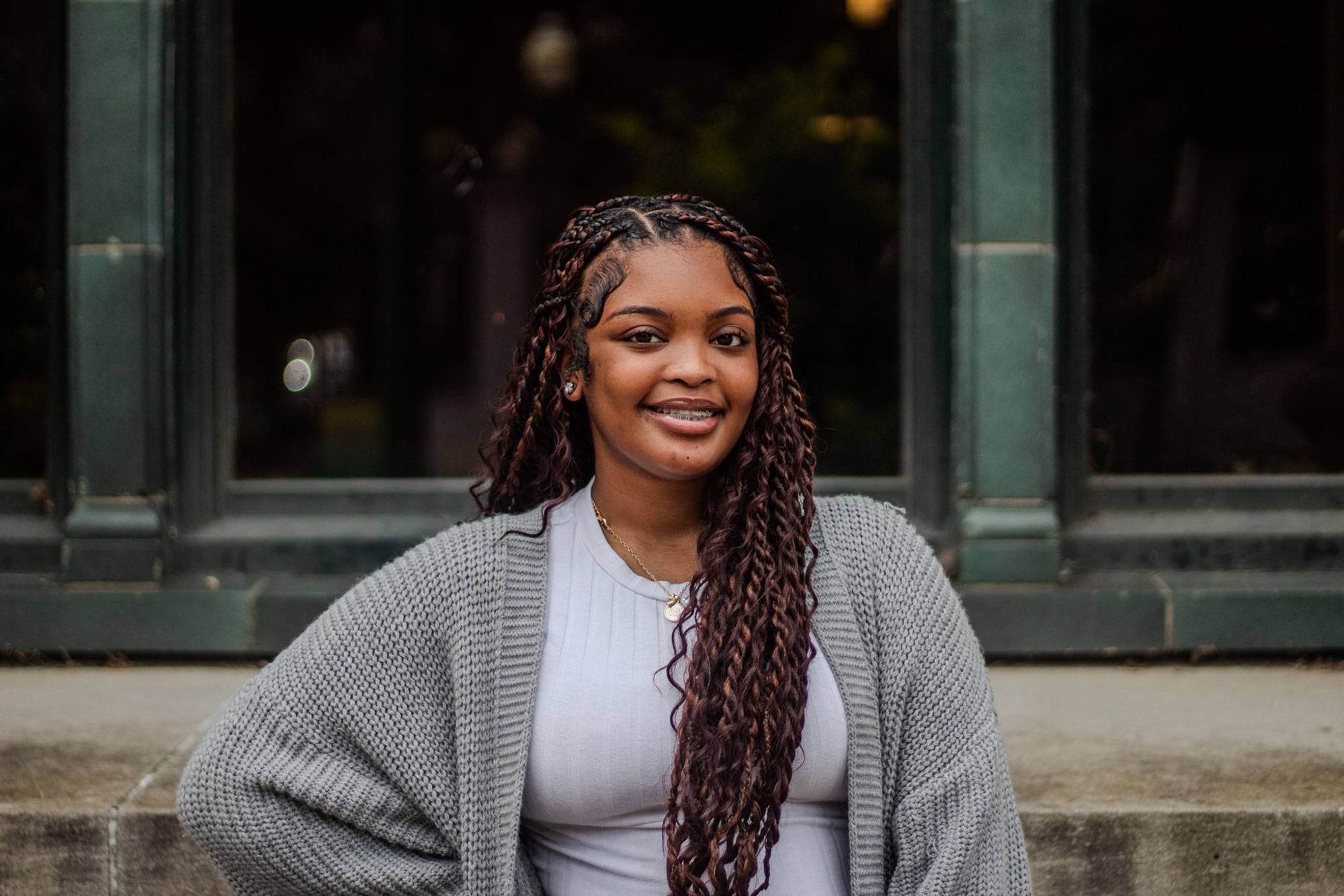 A girl with braids wearing a grey sweater and a white t shirt stands in front of a building with green framing on the outside.
