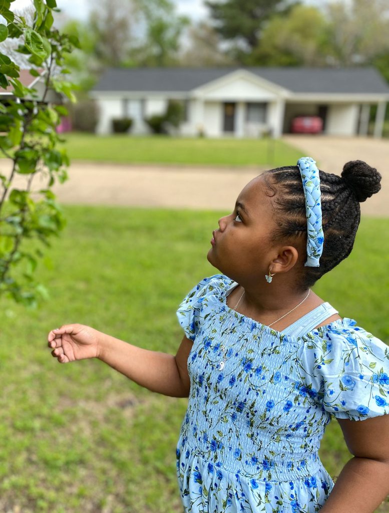 A little girl in a blue dress looks into a bush in the front yard