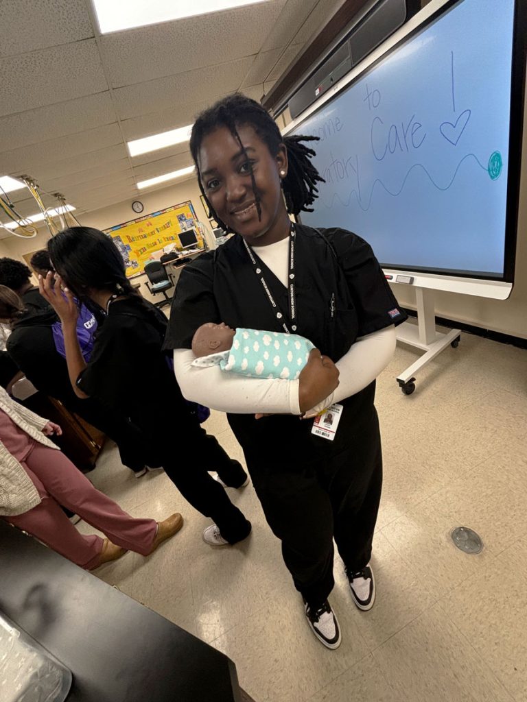 A teen in black healthcare scrubs holds a infant doll to simulate child care