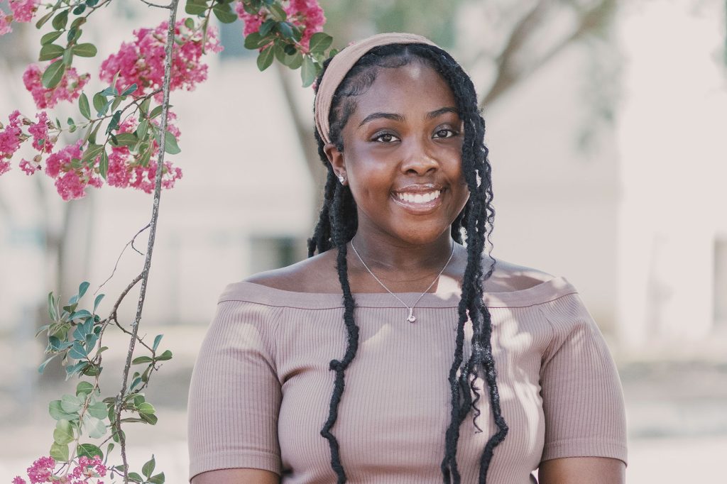 A teen wearing a brown top and matching headband stands outside beside a pink floral vine