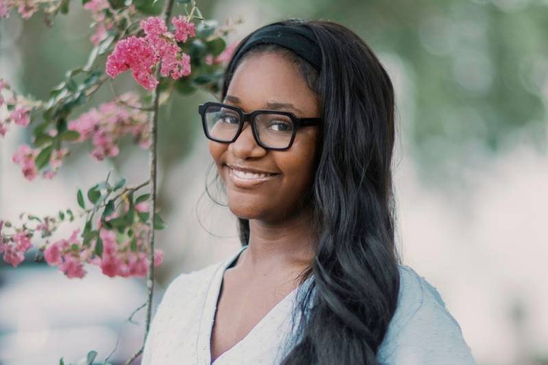 A teen wearing black glasses stands outside beside a pink floral vine