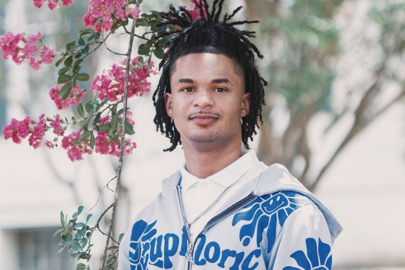 A teen wearing a blue and grey graphic hoodie stands outside beside a pink floral vine