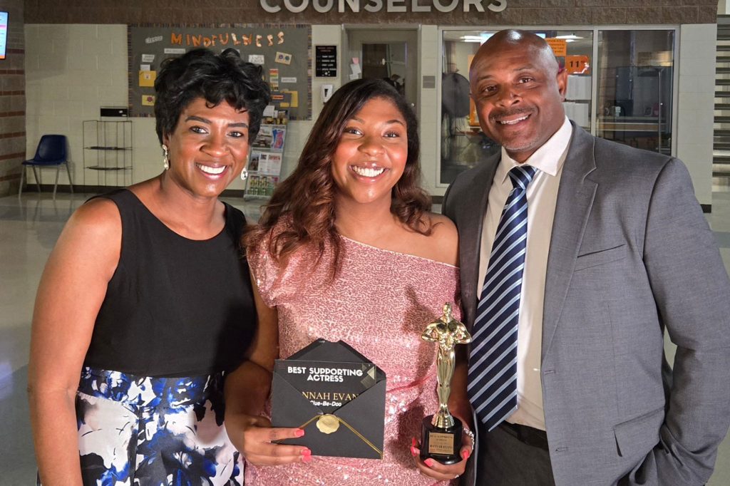 A teen standing between her parents holding a trophy and a card that reads Best Supporting Actress
