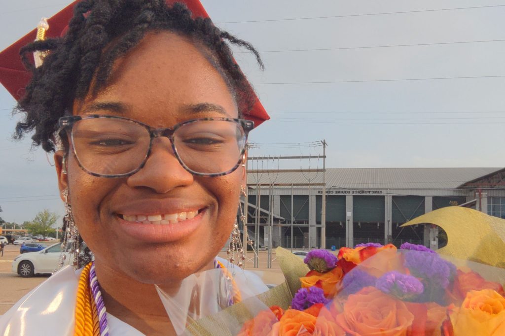 A student in maroon graduation cap poses outside with colorful flowers