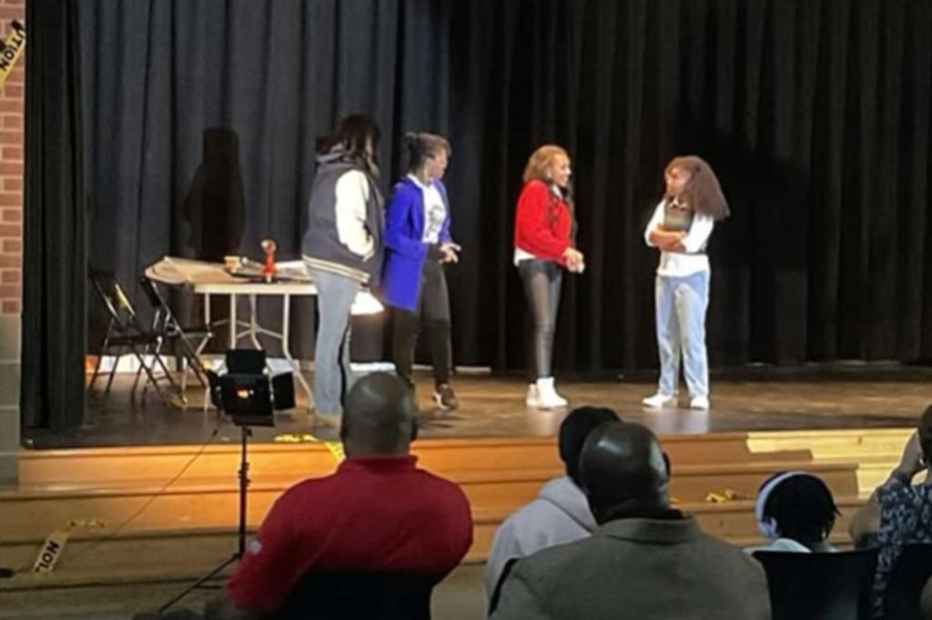Four students stand on stage, acting out a play with limited set decorations