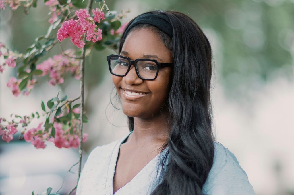 A teen wearing black glasses stands outside beside a pink floral vine