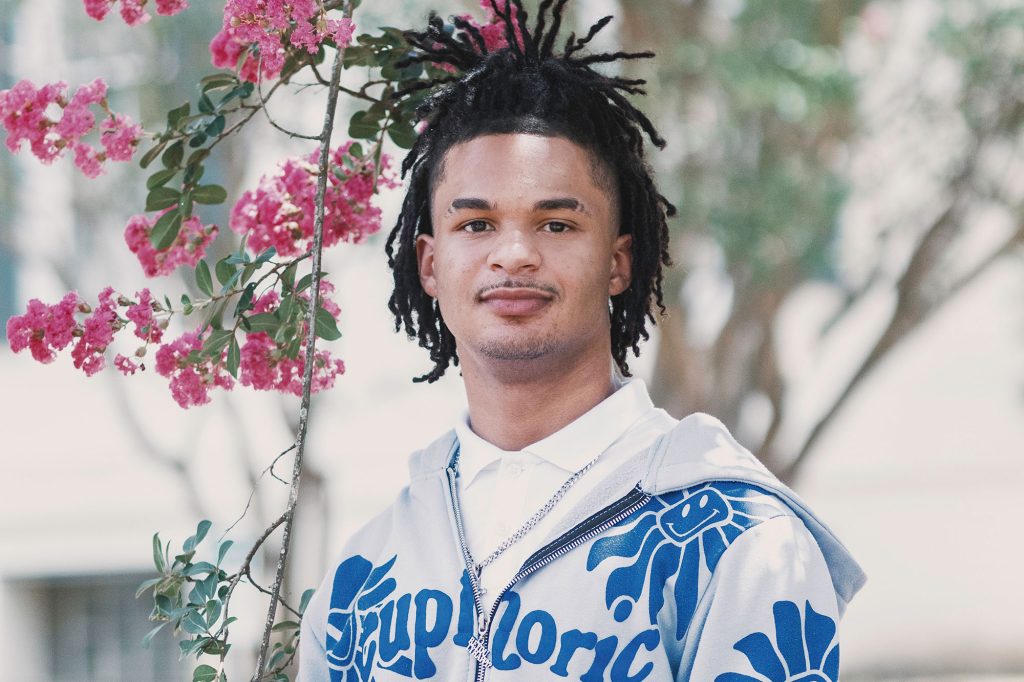 A teen wearing a blue and grey graphic hoodie stands outside beside a pink floral vine