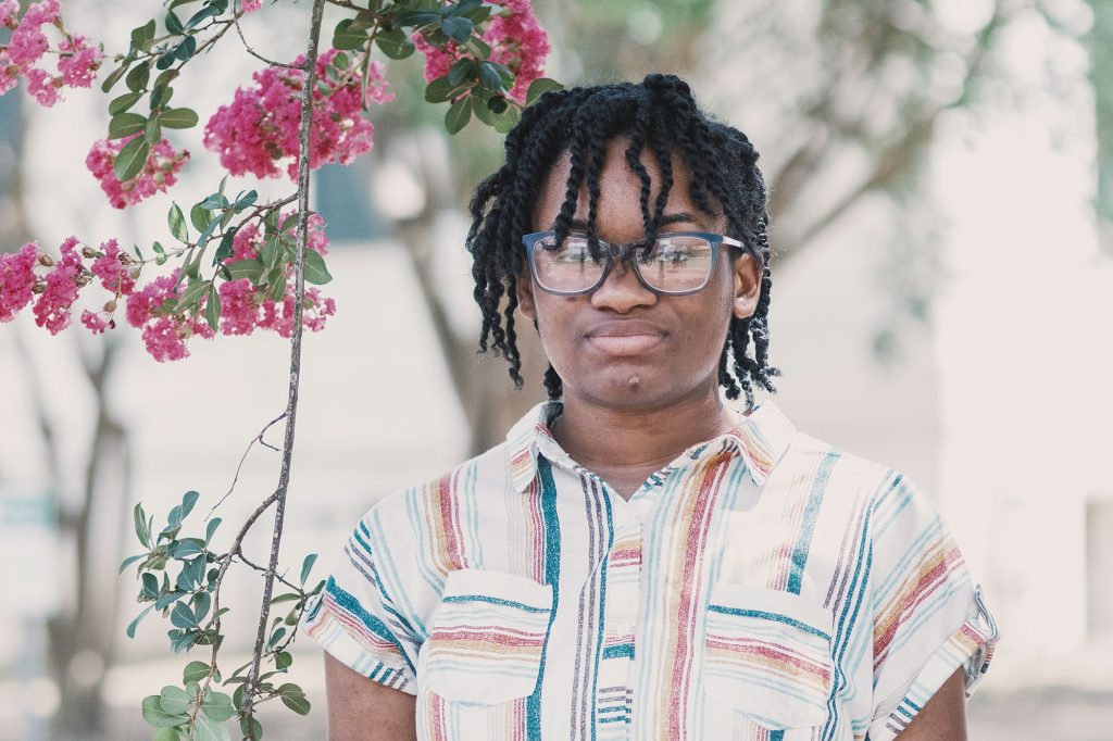 A teen wearing a graphic striped top and glasses stands outside beside a pink floral vine