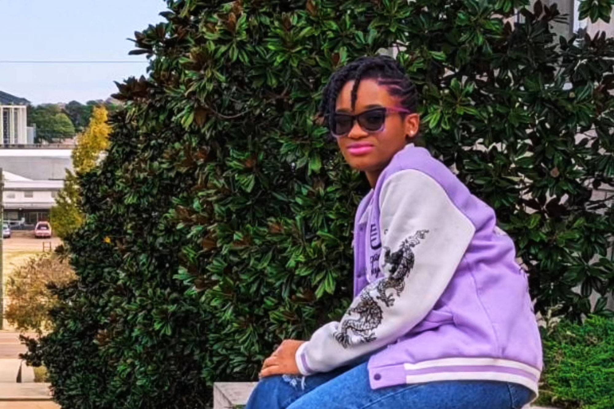 A teen poses outside in a lilac sports jacket and matching sunglasses