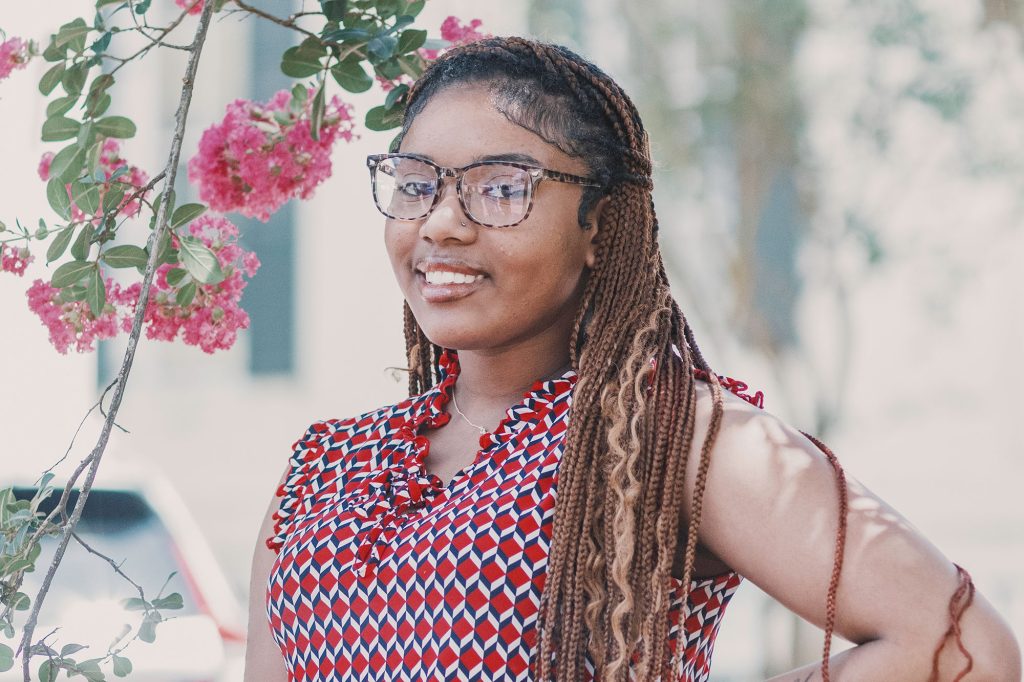 A teen wearing a red graphic patterned top and glasses stands outside beside a pink floral vine
