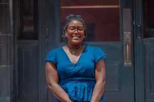 A teen in a blue outfit stands outside in front of green doors