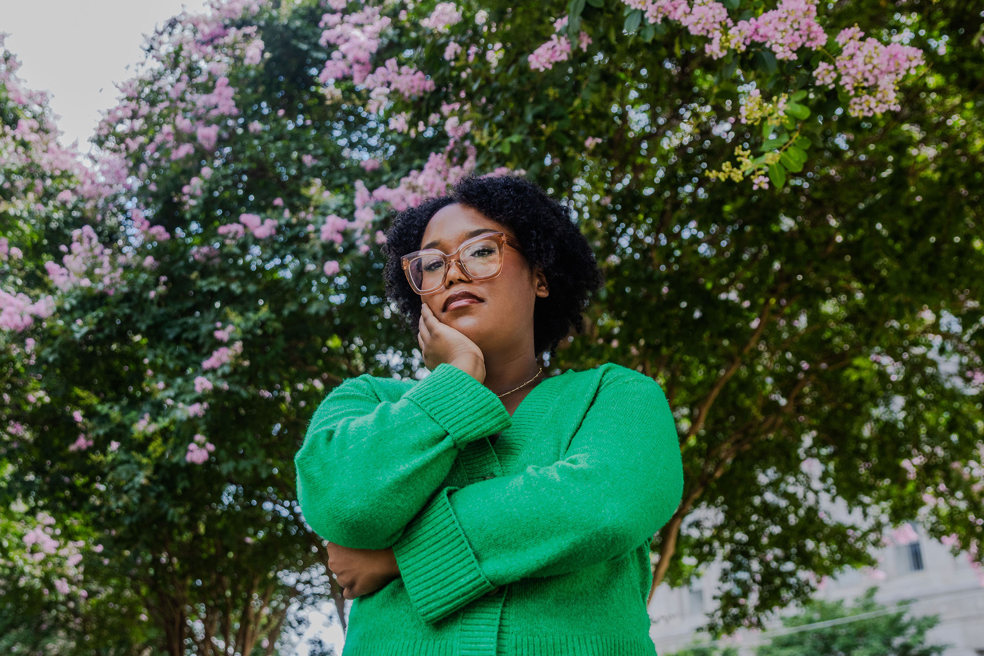 A teen in a green sweater and glasses stands with arms crossed, one hand on chin, in front of a flowering tree