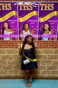 A teen with a yellow sash poses with victory signs in front of campaign election signs for Junior Maid
