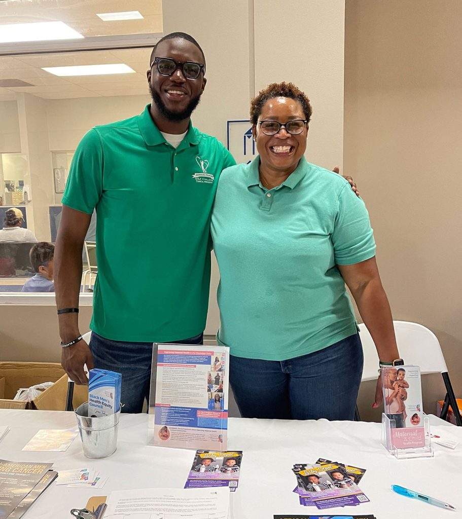 Black man and woman wearing green shirts and standing behind a white table.