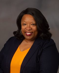 A professional headshot of a smiling woman wearing a black jacket over a gold top