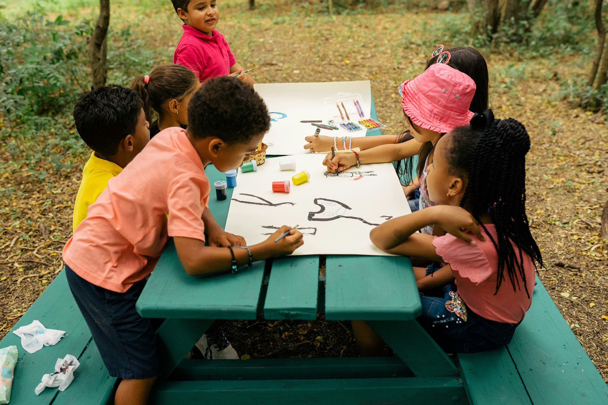 Seven young children sit on a green park bench outside and paint on posterboard