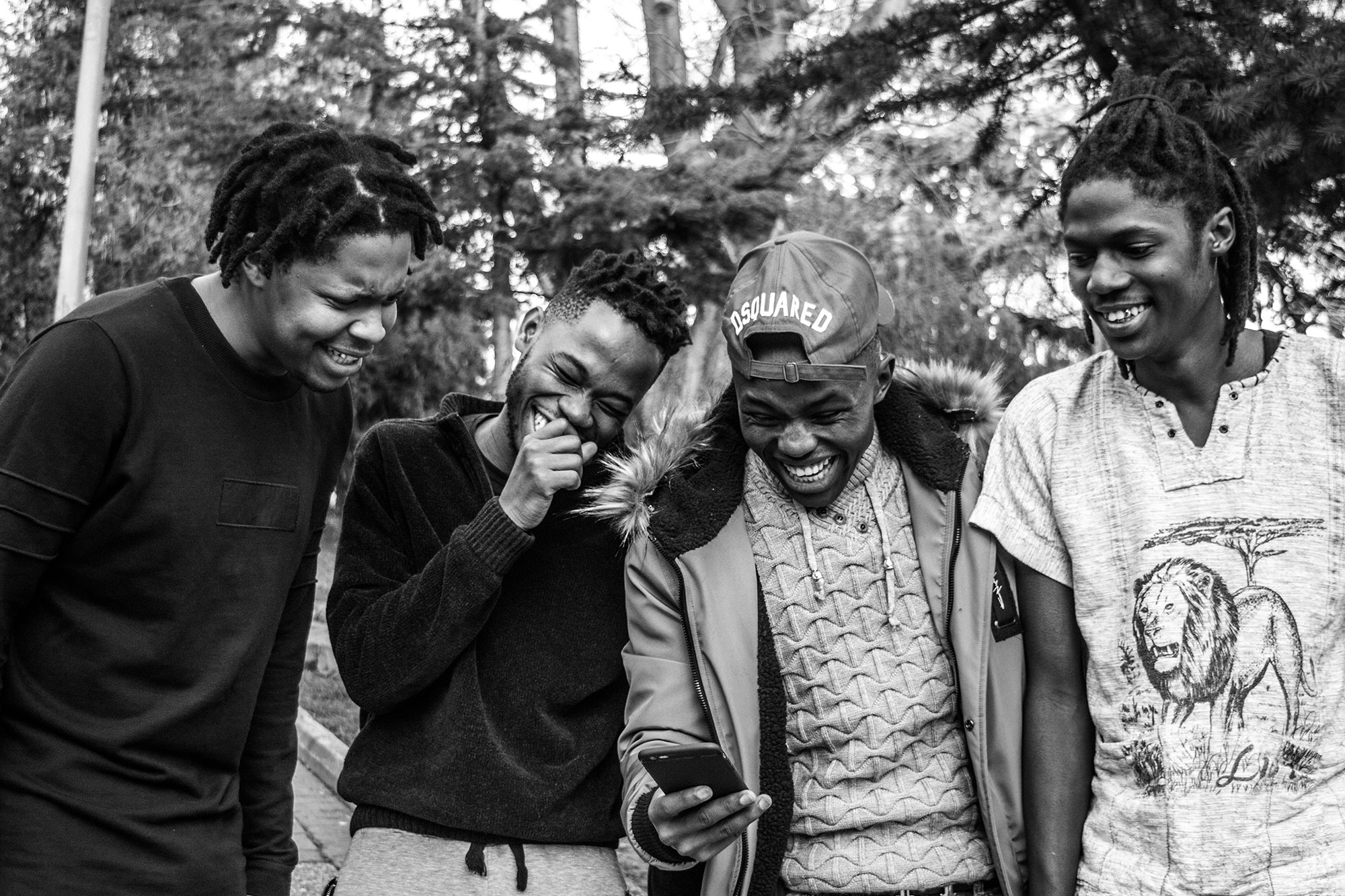 Four Black men looking at a phone, laughing standing outside.