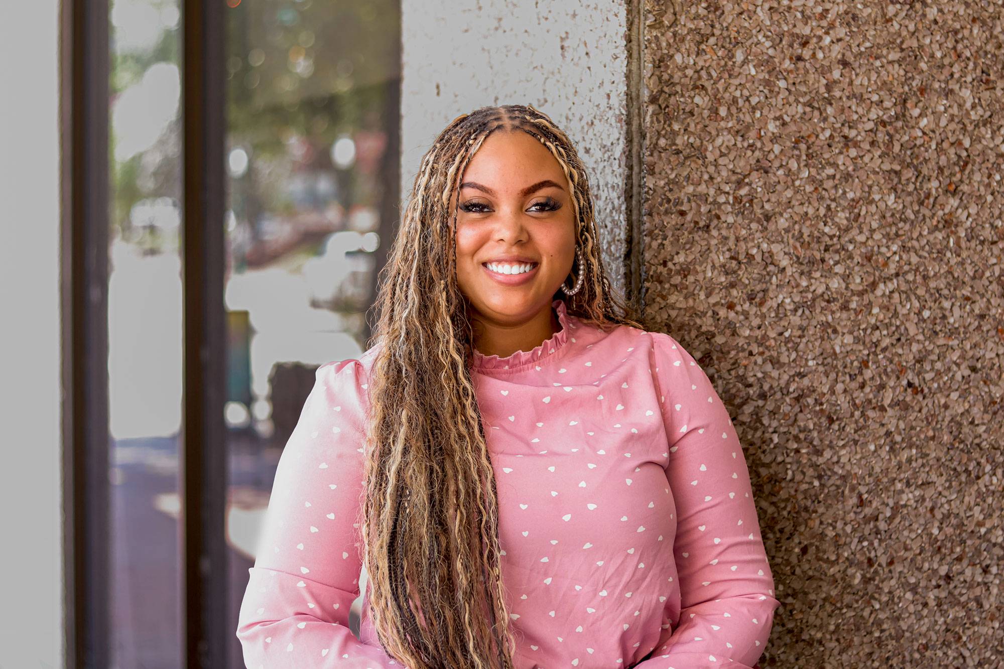 Girl with blonde braids and a pink shirt with white hearts smiling for a picture outside a building.