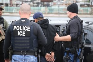 A behind view of two ICE officers escorting a man in handcuffs to a cop car