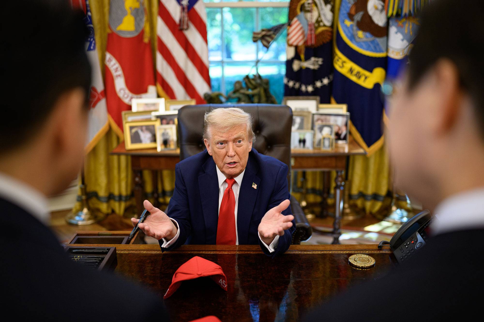Donald Trump speaking at his desk in the Oval Office, as seen over the shoulders of two men