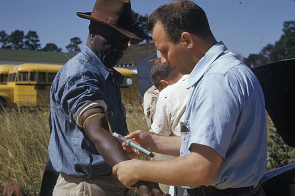 A Black man is being injected with a syringe by a white doctor.
