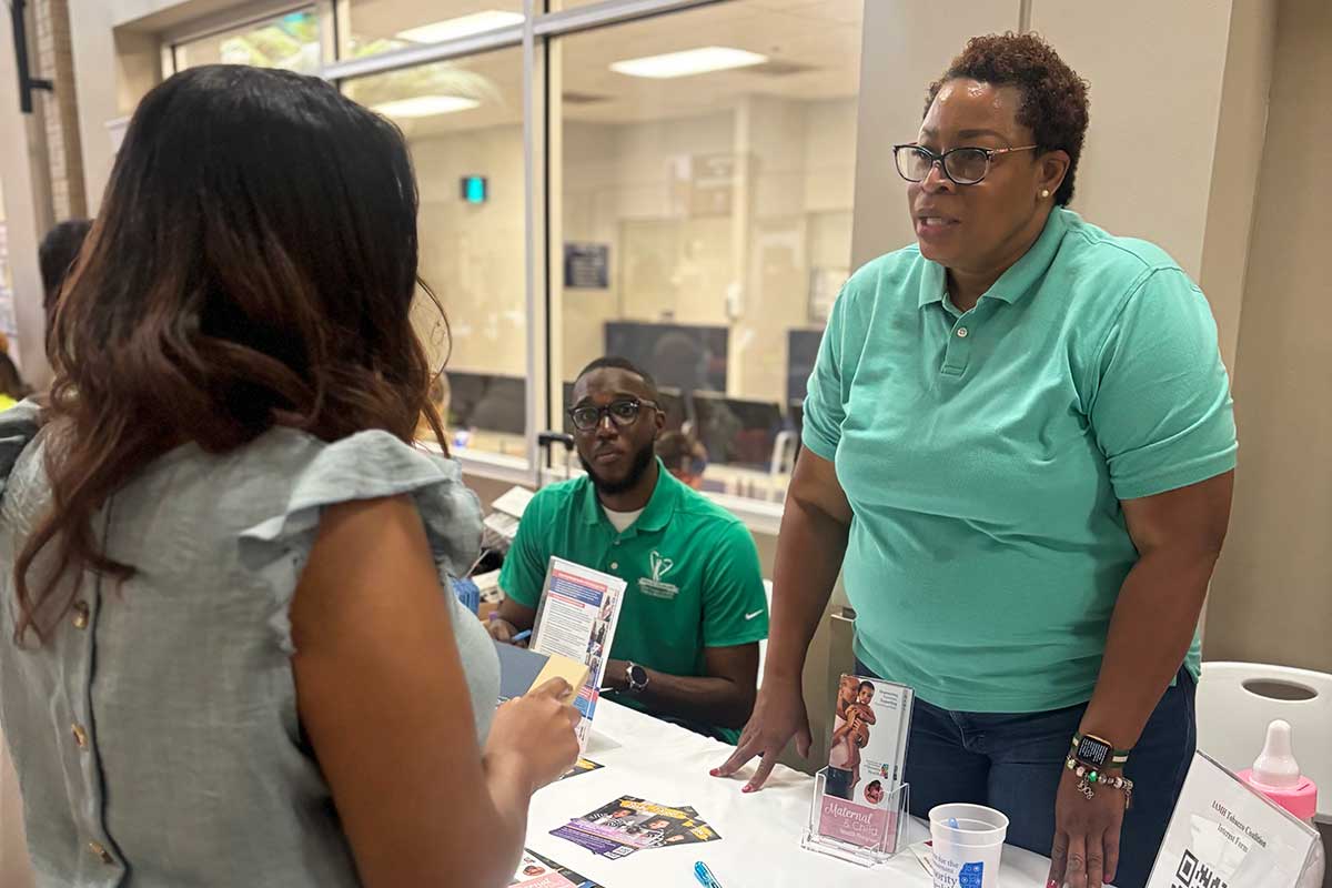 Black girl talking to Black woman with glasses in teal shirt at a table covered in pamphlets